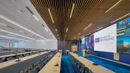 Photo of a large open lecture hall with rows of tables and chairs and a large 48-foot LED screen along one wall for learning.